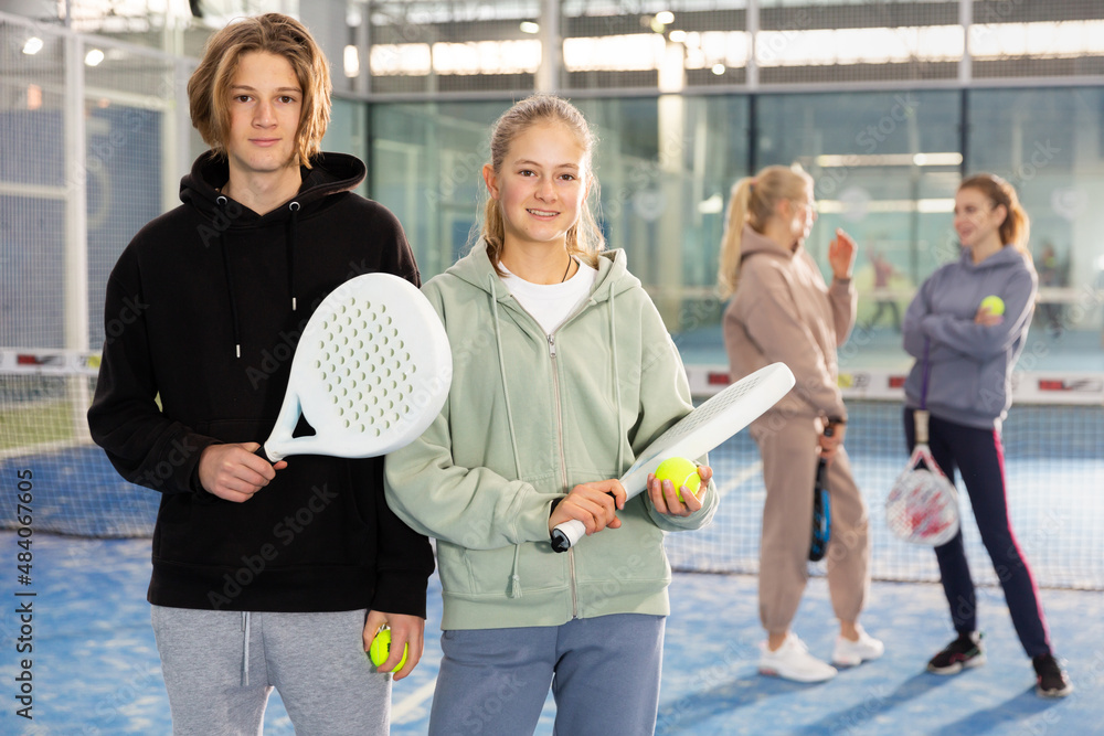 Teenage boy and girl with racquets and balls standing in padel court, looking at camera and smiling.