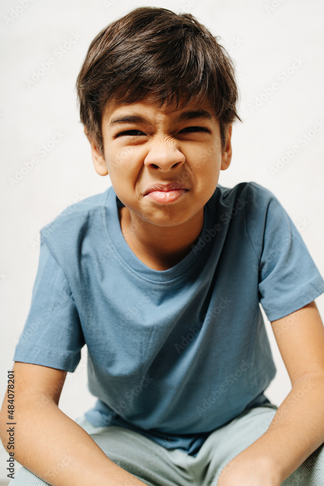 Grimacing indian boy sitting with wrinkled tense face over white background Grimacing indian boy sitting with wrinkled tense face over white background