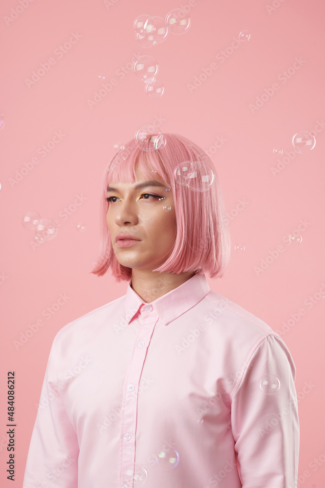 Serious young man in light pink wig standing under soap bubbles and looking away Serious young man in light pink wig standing under soap bubbles and looking away