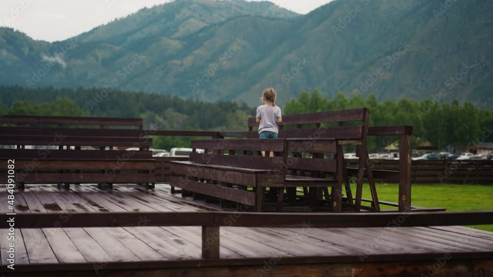 Curious little girl walks along wet wooden benches on large empty dancing ground against old slightly sloping mountains backside view Curious little girl walks along wet wooden benches on large empty dancing ground against old slightly sloping mountains backside view