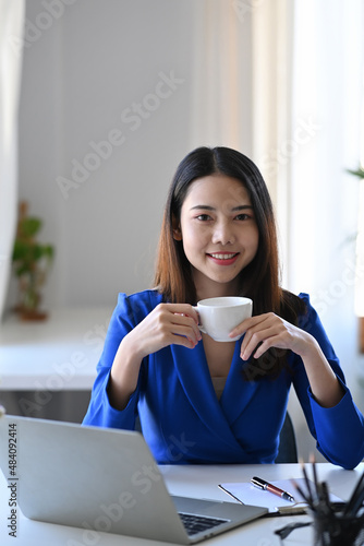 Positive young businesswoman sitting at her workplace and drinking coffee.
