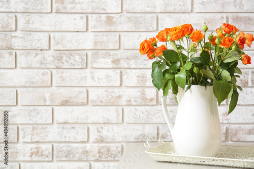 Jug with beautiful orange roses on table against brick wall