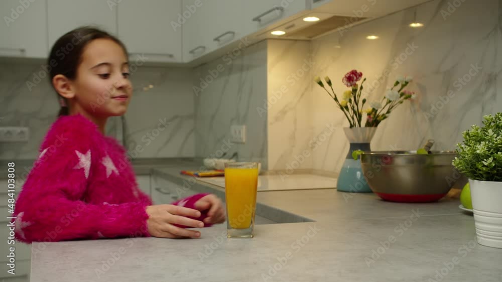 Moving shot of glass of juice standing on kitchen counter while lovely girl approaching table, taking cup, drinking fresh juice indoors. Female child enjoying taste of vitamin drink, smiling at home Moving shot of glass of juice standing on kitchen counter while lovely girl approaching table, taking cup, drinking fresh juice indoors. Female child enjoying taste of vitamin drink, smiling at home