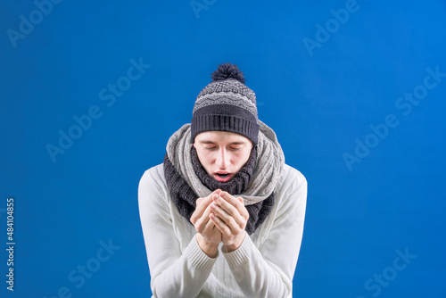 A man in a fur hat and scarf blows warmth on his hands on a blue background. Freezing person. Warmly. Winter. Coldly