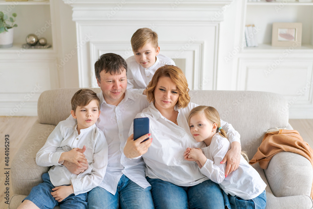 Happy cheerful caucasian family dad mom and kids having fun and using digital phone video call on couch at home. Self-isolation sitting at home social distancing quarantine for coronavirus prevention.