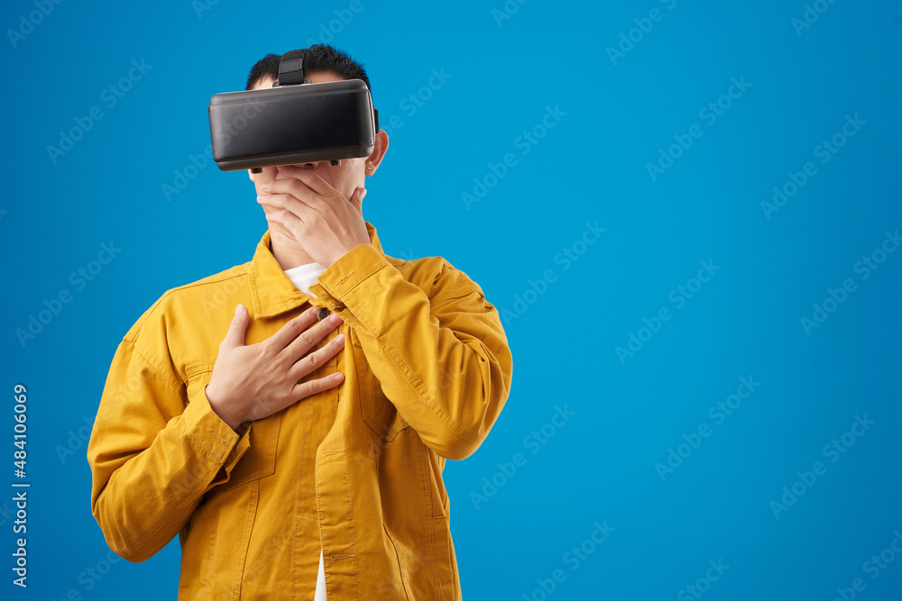 Shocked on scared young man in virtual reality headset covering mouth with hand when looking at something disgusting in videogame Shocked on scared young man in virtual reality headset covering mouth with hand when looking at something disgusting in videogame