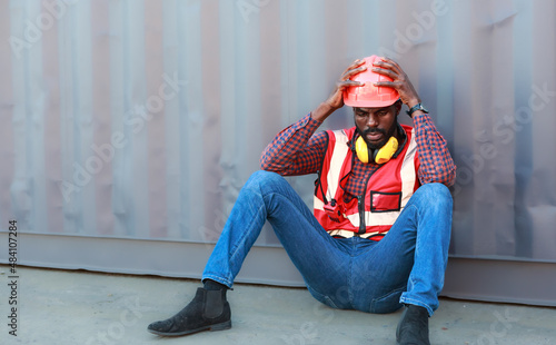 Portrait of an African American male technician or engineer. Sit near a shipping container and look tired and sleepy or unemployed. © atitaph
