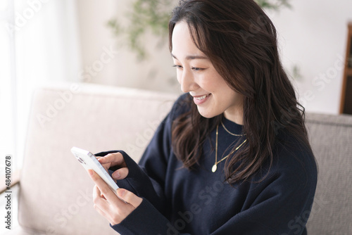 ソファーに座りながら携帯電話を見るアジア人女性、自宅リビング