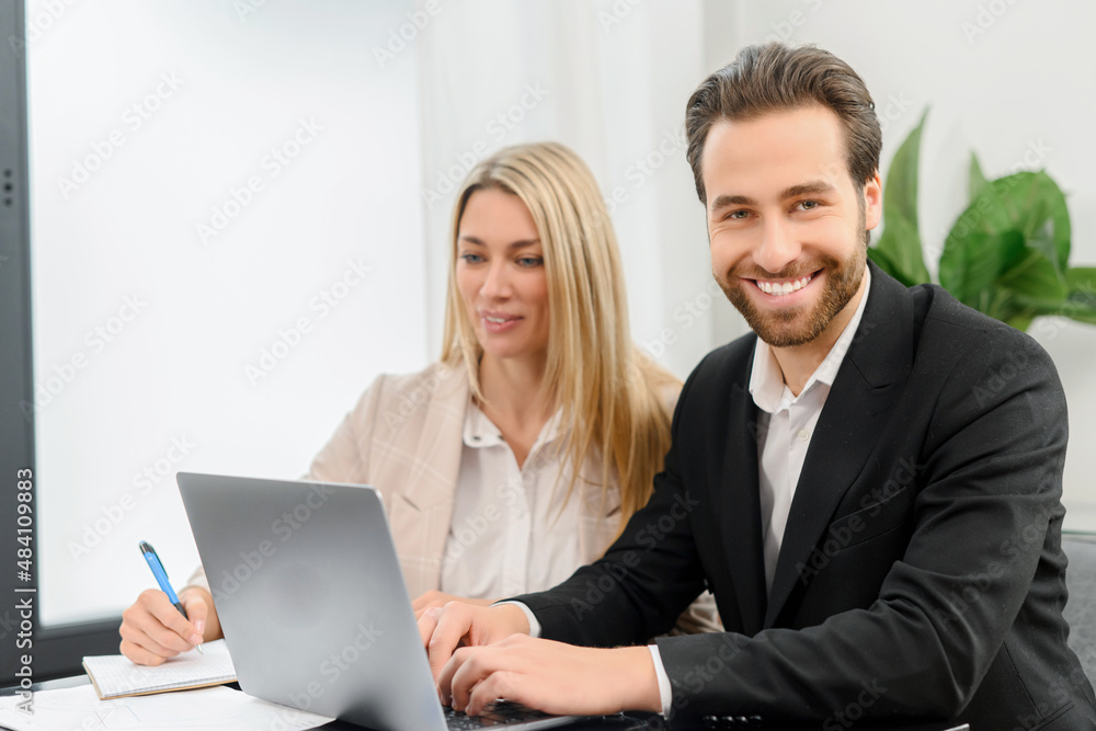 Young attractive man and woman working with papers and laptop in modern office. Blonde girl making notes on paper documents while working with a laptop Young attractive man and woman working with papers and laptop in modern office. Blonde girl making notes on paper documents while working with a laptop