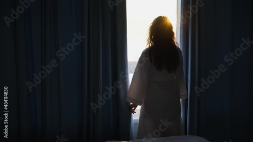 Silhouette woman in white bathrobe opens curtains in dark badroom after waking up looking outside large panoramic window enjoying sunrise in early morning
