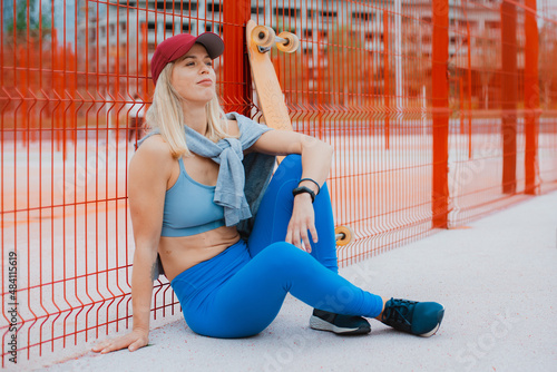 Image of fit perky happy middle aged woman in blue leggings and red cap sitting with skate outside