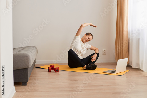 Horizontal shot of athletic woman sitting on mat and using laptop for online workout, wearing white T-shirt and black leggins, stretching her body, workout at home on floor.