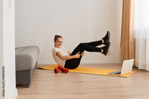 Fitness girl with hair bun wearing white T-shirt and black leggins ,doing sport, physical training on gym mat, reaching hands to feet, exercising abdominal muscles, having fun and laughing.