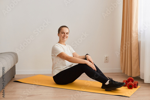 Indoor shot of slim smiling female with hair bun wearing white T-shirt and black leggins, sitting on floor on yoga mat, looking at camera with happy expression, doing sport.