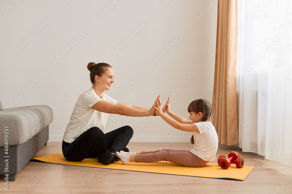 Image of attractive woman mother with hair bun doing sport with her little daughter, sitting on yoga mat on floor and touching each other palms, sport together at home.