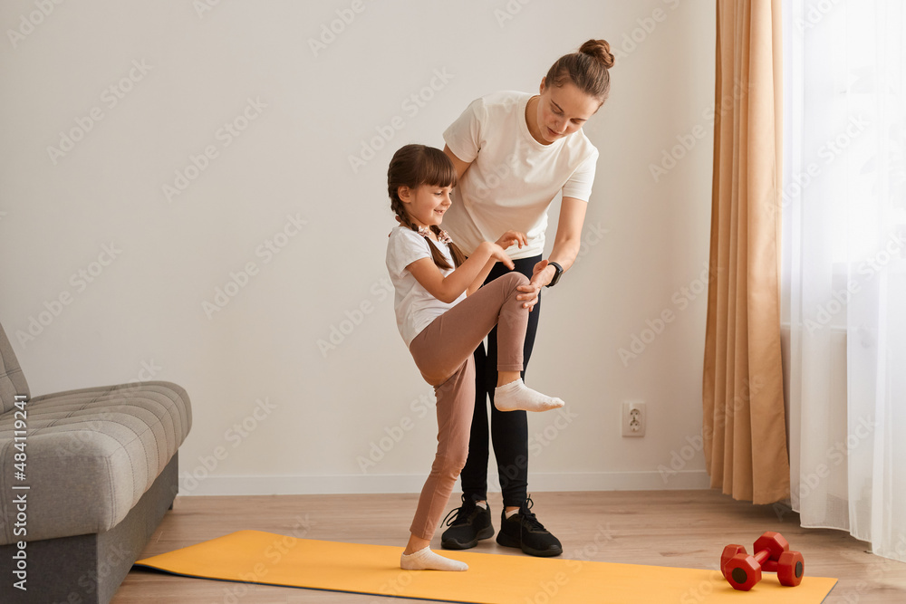 Indoor shot of of female sport trainer teaching little girl to do fitness exercises, kid standing on one leg and stretching, people posing in light room on mat. Indoor shot of of female sport trainer teaching little girl to do fitness exercises, kid standing on one leg and stretching, people posing in light room on mat.