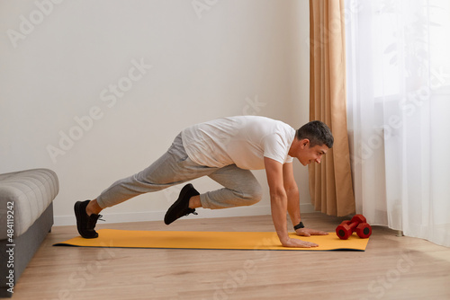 Sport, fitness and healthy lifestyle. Young strong attractive man doing running plank exercise at home near sofa and window, wearing sportswear and sneakers.
