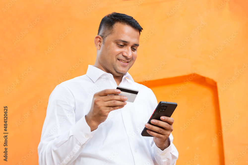 Online banking and payment concept. Smiling indian man using credit or debit card while holding smart phone against orange background. Satisfied asian guy shopping, buying on internet. happy customer Online banking and payment concept. Smiling indian man using credit or debit card while holding smart phone against orange background. Satisfied asian guy shopping, buying on internet. happy customer