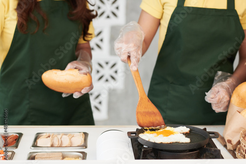 Cropped image of vendors in aprons making sandwich with meat and fried egg for customer