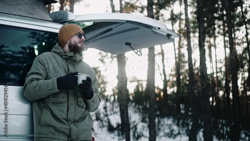 Man drinks leaning on the back of a van with pop up roof in the woods in winter. Tourist living in a van with pop-up roof. Camper having drink at the woods. High quality 4k footage