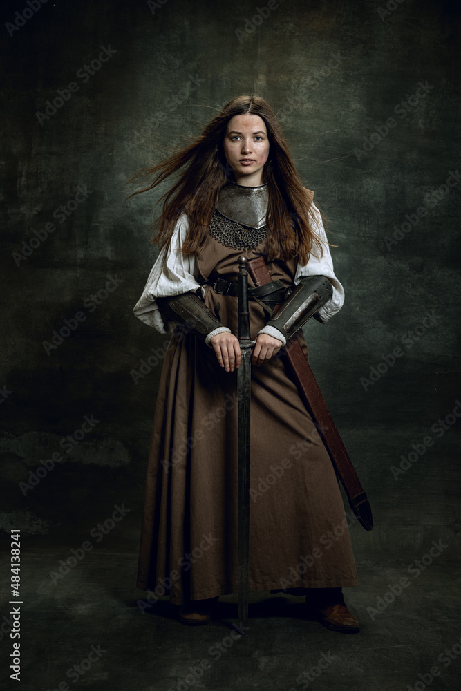 Full-length portrait of seriuos beautiful woman in image of medieval warrior or knight with dirty wounded face with big sword isolated over dark vintage background. Full-length portrait of seriuos beautiful woman in image of medieval warrior or knight with dirty wounded face with big sword isolated over dark vintage background.