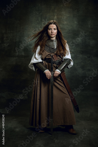 Full-length portrait of seriuos beautiful woman in image of medieval warrior or knight with dirty wounded face with big sword isolated over dark vintage background.