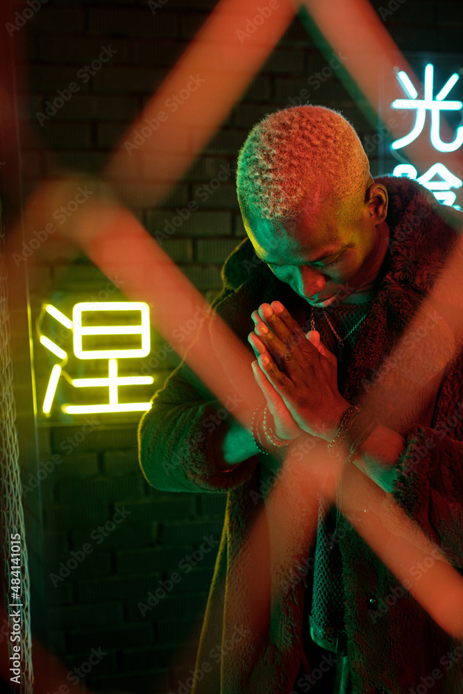 Obraz premium Young African man in furcoat keeping his hands put together and bowing while standing behind bars against illuminated hieroglyphs