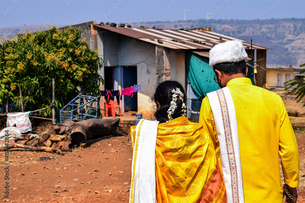 Fototapeta premium Back view of newly married Indian couple, bride wearing yellow color saree ,groom wearing yellow color kurta and white color cap. Both are walking toward their home