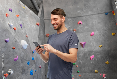 fitness, sport and bouldering concept - smiling man in sports clothes with smartphone and earphones listening to music over climbing wall at gym on background