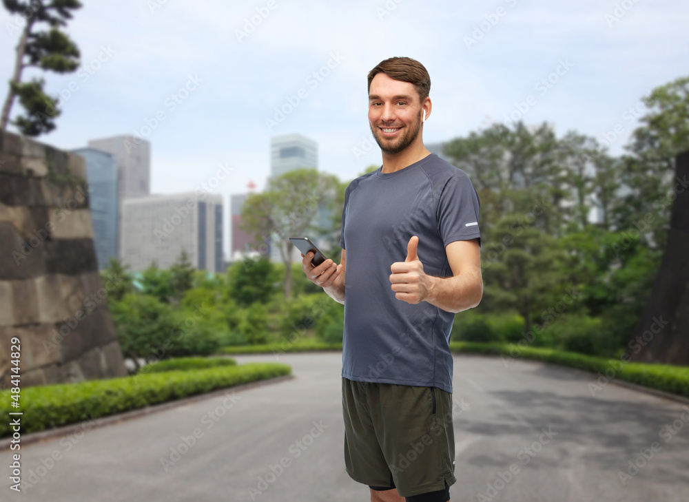 Fototapeta premium fitness, sport and healthy lifestyle concept - smiling man in sports clothes with smartphone and earphones listening to music and showing thumbs up gesture over park in tokyo city, japan on background