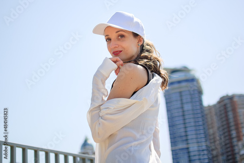 Female portrait of young active girl in black top, white shirt, basketball cap, and jeans on modern buildings background outdoors