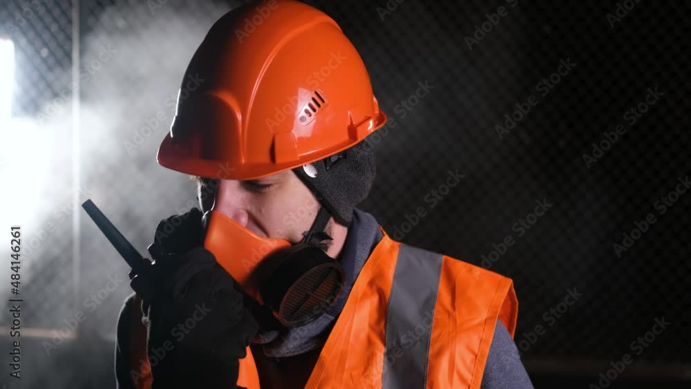 The camera moves left-right. A worker in an orange helmet and overalls shouts into a walkie-talkie, reporting a disaster at work. Smoke rises behind him The camera moves left-right. A worker in an orange helmet and overalls shouts into a walkie-talkie, reporting a disaster at work. Smoke rises behind him