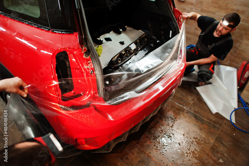 Car service worker put anti gravel film on a red car body at the detailing vehicle workshop. Car protection with special films.