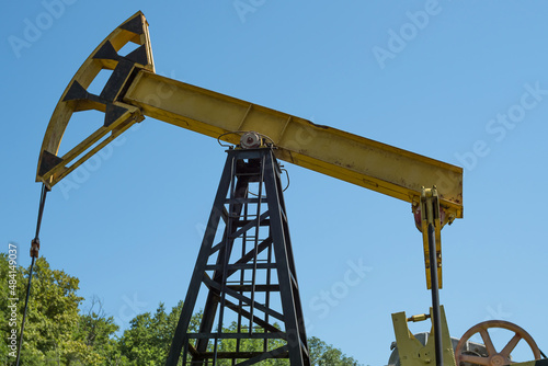 Close-up of an old oil rig pumping oil against a backdrop of green trees and blue sky. Side view from below. Extraction of black gold from the well.