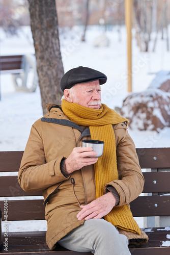 Elderly man in warm clothing sitting on the bench and drinking hot tea, he resting outdoors in the park in winter