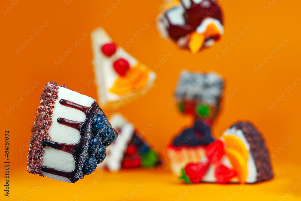 Various cakes fall on an orange background. Glazed triangular pieces of cake with chocolate filling and fruit creams. Sweets fly in the air.