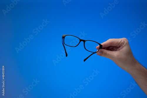 Black plastic frame clear glass eyeglasses held by male hand. Close up studio shot, isolated on blue background