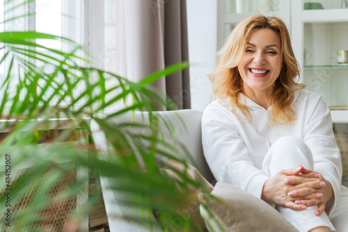 Indoor portrait of attractive European woman in her 40 plus with blonde hair and fit body looking at camera with pleased friendly smile, sitting on comfortable gray couch photo