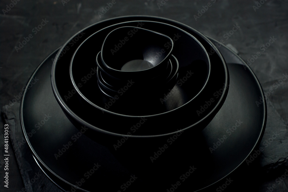 Black tableware on a dark background. Plates and bowls of different shapes and sizes are stacked in a pile. Contrasting dramatic light as an artistic effect.