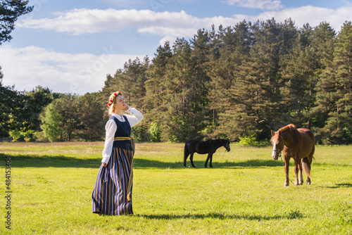 Latvian woman in traditional clothing posing on nature with horse in village.