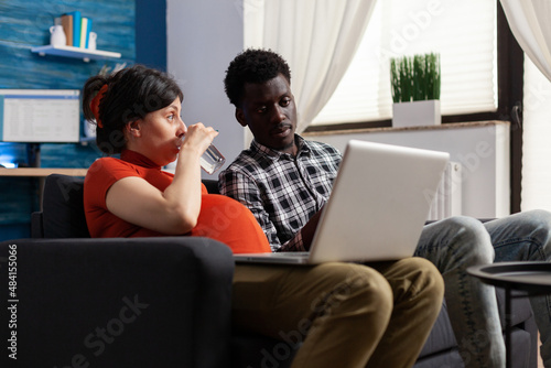 African american man looking at laptop while pregnant caucasian woman is drinking a glass of water on sofa. Interracial couple sitting together in living room while future mother drinking water with