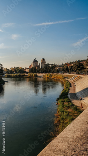 Photo taken over Ponte Pietra in Verona, Italy. In the background there are houses, and a basilica on top of the mountain, with a blue sky and a bird flying.