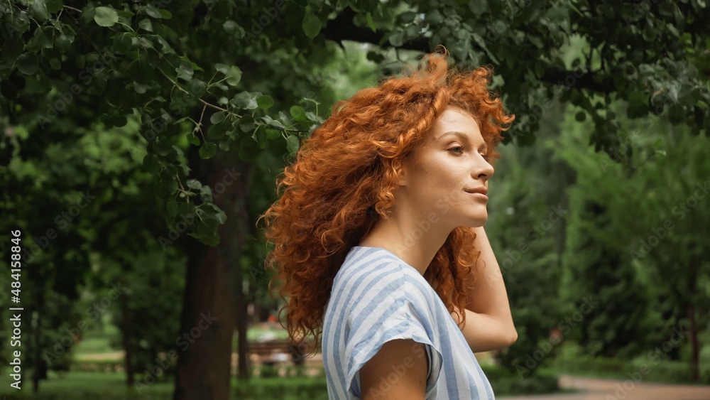 Naklejka premium curly young woman with red hair standing in green park.