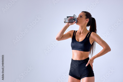 athletic young woman drinking fresh water after active training, holds water bottle, dressed in black sportswear, isolated over gray concrete wall. People and weariness concept. portrait © alfa27