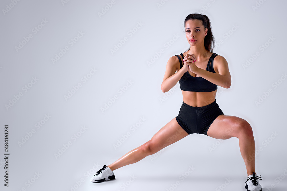 young caucasian woman coach in sporty black short top and gym shorts makes lunges by the feet side, hands held together, exercising on white isolated background in studio. sport and fitness concept