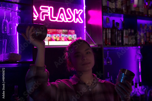 Young smiling caucasian woman bartender with cocktail shaker in nightclub bar with red pink and violet purple neon light, barman looking at camera at club party, selective focus