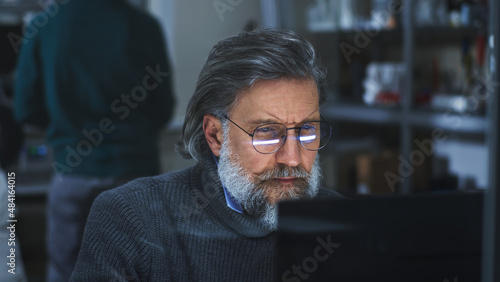 Middle aged bearded man in glasses creating 3D model on computer while sitting behind glass in contemporary printing workshop