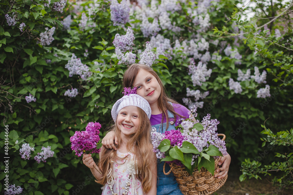 Two smiling European girls stand in a summer garden with a basket of lilacs in their hands