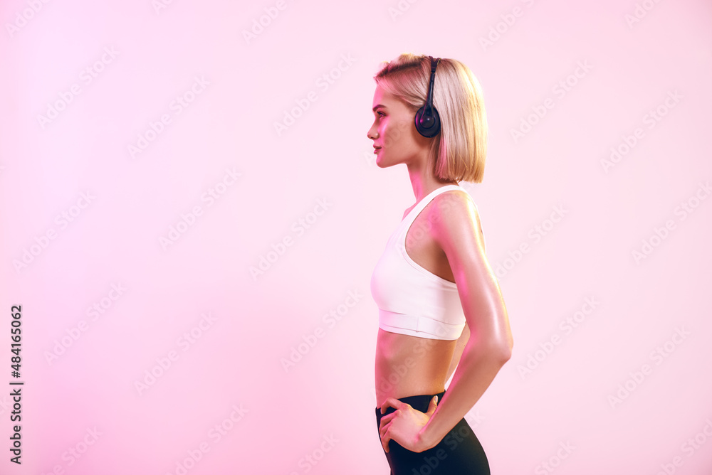 Obraz premium Music in my life. Side view of sporty blonde woman in headphones listening music while standing against pink background