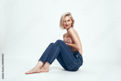 Natural beauty. Full length of cute young woman in jeans covering breasts with hands and looking aside while sitting on the floor against grey background in studio